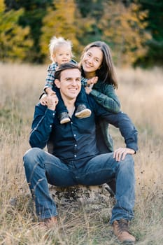 Smiling mom hugging dad from behind with a little girl on his shoulders in a clearing. High quality photo