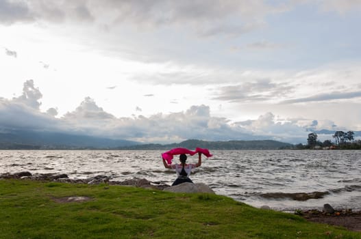 indigenous girl sitting on the shore of a lagoon with her arms raised and a fuchsia veil looking towards the horizon where there are many clouds. earth day. High quality photo