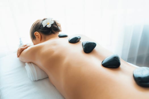 Hot stone massage at spa salon in luxury resort with day light serenity ambient, blissful woman customer enjoying spa basalt stone massage glide over body with soothing warmth. Quiescent