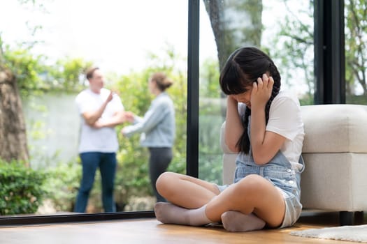 Stressed and unhappy young girl huddle in corner, cover her ears blocking sound of her parent arguing in background. Domestic violence at home and traumatic childhood develop to depression. Synchronos