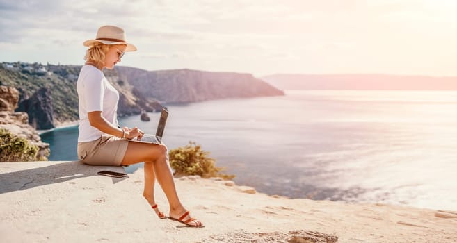 Successful business woman in yellow hat working on laptop by the sea. Pretty lady typing on computer at summer day outdoors. Freelance, travel and holidays concept.