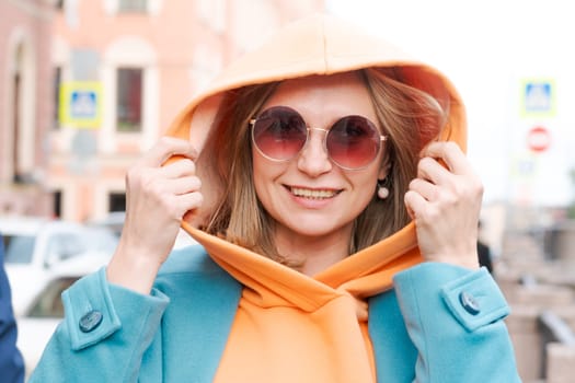 Happy woman walking through city with bag in her hand, dressed in bright clothes, an orange hoodie and a blue coat, smiling contentedly in sunglasses