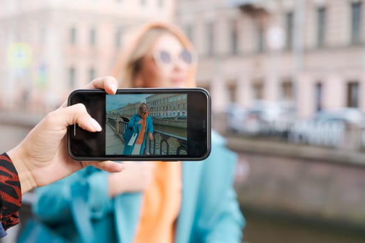 Woman posing on phone near canal dressed in bright clothes orange hoodie and blue coat, smiling contentedly, lifestyle concept
