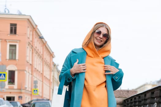 Happy woman walking through city with bag in her hand, dressed in bright clothes, an orange hoodie and a blue coat, smiling contentedly in sunglasses