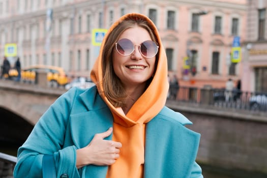 Happy woman walking through city with bag in her hand, dressed in bright clothes, an orange hoodie and a blue coat, smiling contentedly in sunglasses