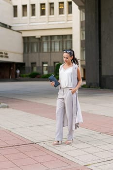 Middle-aged woman dressed in jacket and trousers walks around city against backdrop of a business center with a notepad in her hand