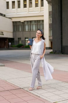 Middle-aged woman dressed in jacket and trousers walks around city against backdrop of a business center with a notepad in her hand