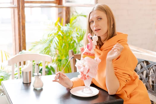 Beautiful woman eats dessert in form pink flamingo in cafe. Time for fun, vows in a bright sweatshirt