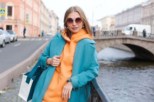 Woman walking through city near canal dressed in bright clothes orange hoodie and blue coat, smiling contentedly, lifestyle concept