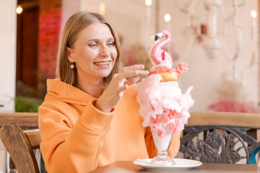 Happy woman eating sweet dessert in form pink flamingo in restaurant, woman sitting in a cafe and enjoying a sweet dessert for lunch, sweet tooth concept