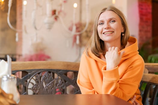 Cute woman in cafe. Portrait beautiful woman, alone at table in cafe. Concept waiting for an order. In a bright orange sweatshirt