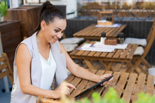 Beautiful businesswoman with smile sitting with touchpad in cozy restaurant while relaxing outside, happy female student working on a digital tablet and relaxing in a cafe after university