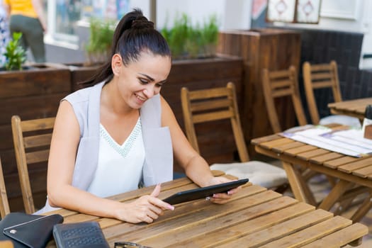 Beautiful businesswoman with smile sitting with touchpad in cozy restaurant while relaxing outside, happy female student working on a digital tablet and relaxing in a cafe after university