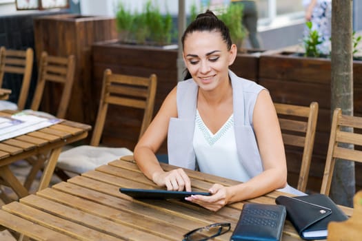 Beautiful businesswoman with smile sitting with touchpad in cozy restaurant while relaxing outside, happy female student working on a digital tablet and relaxing in a cafe after university