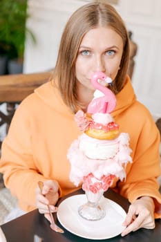 Beautiful woman eats dessert in form pink flamingo in cafe. Time for fun, vows in a bright sweatshirt