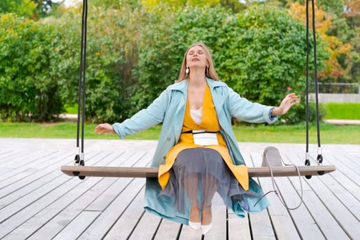 Happy woman swinging on swing in city park, wearing yellow dress and blue coat, the concept of freedom from worries and problems