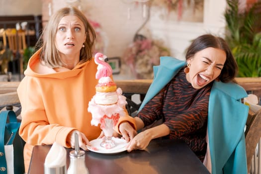 Two women cannot share dessert in form pink flamingo in cafe, they push plate in different directions and laugh, the concept of a sweet tooth