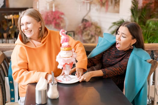 Two women cannot share dessert in form pink flamingo in cafe, they push plate in different directions and laugh, the concept of a sweet tooth