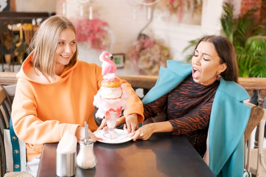 Two women cannot share dessert in form pink flamingo in cafe, they push plate in different directions and laugh, the concept of a sweet tooth