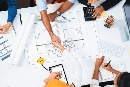 Top panorama of diverse group of civil engineer and client shake hand after make successful agreement on architectural project, review construction plan and building blueprint at meeting table.Prudent