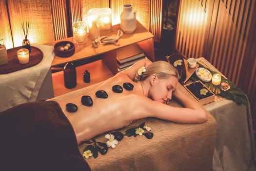 Hot stone massage at spa salon in luxury resort with warm candle light, blissful woman customer enjoying spa basalt stone massage glide over body with soothing warmth. Quiescent