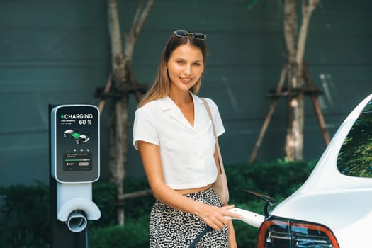 Young woman travel with EV electric car charging in green sustainable city outdoor garden in summer. Urban sustainability lifestyle by green clean rechargeable energy of electric BEV vehicle innards