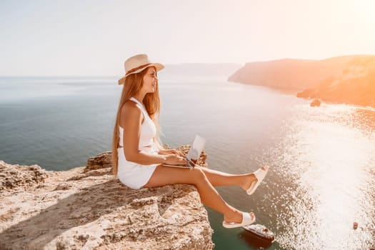 Successful business woman in yellow hat working on laptop by the sea. Pretty lady typing on computer at summer day outdoors. Freelance, travel and holidays concept.