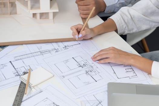 Top view image of professional beautiful young architect hand drafts blueprint while asian cooperative coworker measure house model on table with house model and blueprint scatter around. Immaculate.