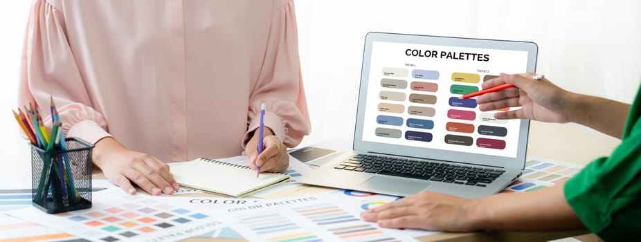 Creative graphic design presents selective color while manager using laptop comparing suitable color at table with color palettes, graphic material scatter around at modern office. Variegated.