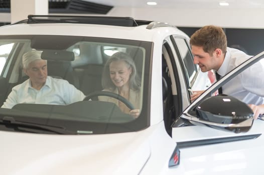 A gray-haired Caucasian woman is sitting behind the wheel of a car. Car dealership salesman consulting mature married couple