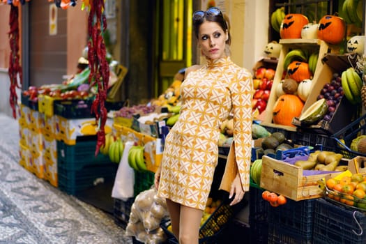 Positive young female in casual yellow gown and makeup, standing and leaning back towards stall with assorted fresh ripe fruits in local market at town in daylight