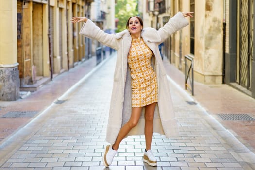 Stylish young woman wearing fashionable long coat with dress and shoe dancing on street against building background