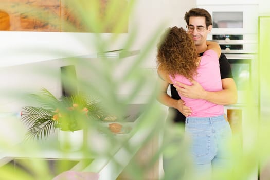 Peek view of smiling young couple in casual clothes embracing while standing near green plants and flowers on terrace of modern apartment