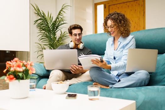 Cheerful young husband and wife in casual clothes sitting on sofa with laptops, while writing on notebook and working together on remote project in cozy living room