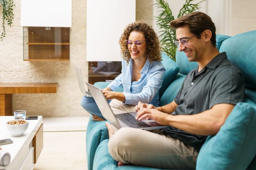 Positive young man and woman in smart casual clothes sitting on sofa smiling and typing on laptop, while looking at screen and working on remote project at home