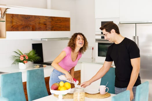 Positive couple having fun while standing near dining table laughing female with eyes closed, pouring juice and joking looking down male in eyeglasses holding glass in kitchen