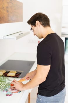 Side view of adult male in casual clothes and eyeglasses looking down while standing in kitchen near cooking range, with bread slices on grill and cutting tomato with knife in daylight