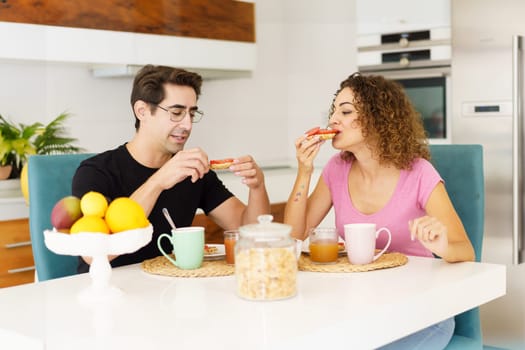 Happy adult couple in conversation and looking down while sitting at dining table in kitchen with juice in glasses, on placemat near fruits in bowl and eating salad in daylight at home