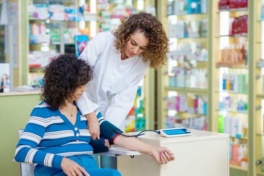Young female pharmacist in white robe, while standing and putting inflatable cuff around sitting lady customer hand during blood pressure measuring near ,shelves with medical supplies in pharmacy