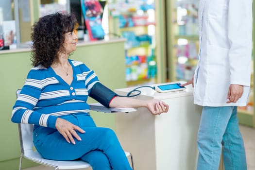 Side view of anonymous female pharmacist in white robe, while standing near legs crossed sitting lady customer during measuring of blood pressure in device in pharmacy