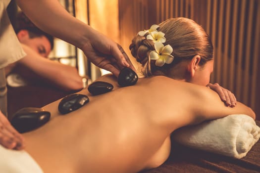 Hot stone massage at spa salon in luxury resort with warm candle light, blissful couple customer enjoying spa basalt stone massage glide over body with soothing warmth. Quiescent