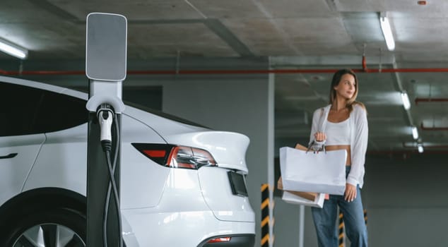 Young woman travel with EV electric car to shopping center parking lot charging in downtown city showing urban sustainability lifestyle by green clean rechargeable energy of electric vehicle innards