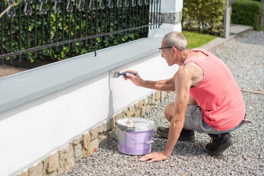 A middle-aged man paints a fence with white paint with a brush on a sunny summer day, repairs the damaged surface, the owner does everything himself, high quality photo