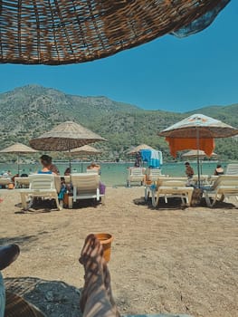 female legs resting in the shade of a wicker beach umbrella on a sandy beach surrounded by mountains. concept of beach holiday, summer vacation. vertical photo