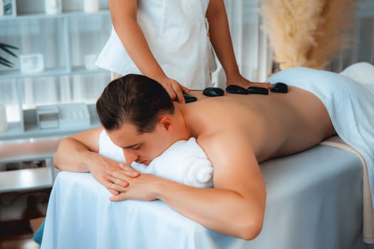 Hot stone massage at spa salon in luxury resort with day light serenity ambient, blissful man customer enjoying spa basalt stone massage glide over body with soothing warmth. Quiescent