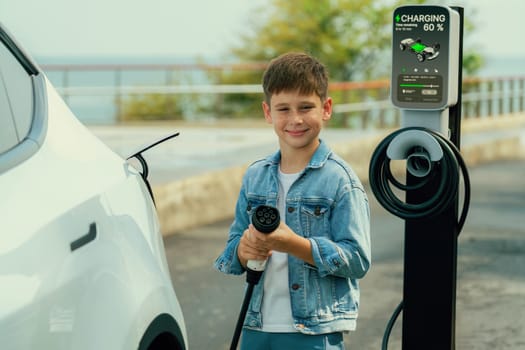 Little boy recharging eco-friendly electric car from EV charging station. EV car road trip travel by the seashore by alternative vehicle powered by clean renewable and sustainable energy. Perpetual