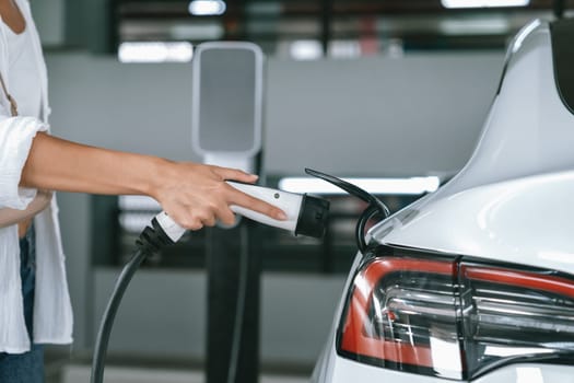 Young woman travel with EV electric car to shopping center parking lot charging in downtown city showing urban sustainability lifestyle by green clean rechargeable energy of electric vehicle innards