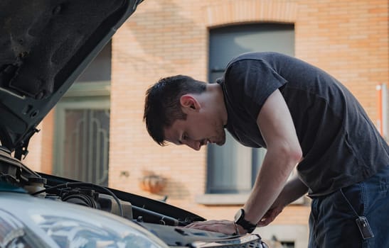 One young caucasian guy looks into the open hood of his car while standing on a city street on a summer afternoon in the evening, close-up side view. The concept of replacing light bulbs, car repair.