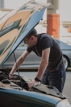 One young caucasian guy takes out a burned-out left headlight bulb from the open hood of his car while standing on a city street on a summer afternoon in the evening, close-up side view. The concept of replacing light bulbs, car repair.