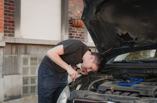 One young caucasian guy bending over unscrews the burnt out bulb of the headlight of his car with a screwdriver, standing on the city street on a summer afternoon in the evening, close-up side view. The concept of replacing light bulbs, car repair.